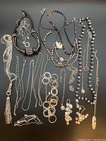 All necklaces arranged on a gray surface showing variety of black and silver colors, chains, beads, pendants, and charm styles.