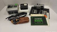 Full lot display showing Olympus 35RC camera, Rollei E15B flash in box, black carrying strap, brown leather case with opera glasses, and paperwork including manuals.