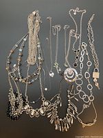 Multiple silver colored necklaces laid flat on dark background showing various designs and pendants.