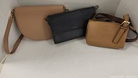 Three ladies purses in neutral colors including a light camel saddle-shaped purse, a black slim rectangular purse, and a tan structured purse with a thick strap