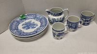 Six blue and white Spode Blue Room dinner plates stacked with a water pitcher and 3 mugs beside them.