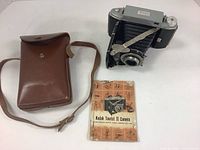 Kodak Tourist II camera with original brown leather case and instruction manual laid next to each other on white surface.