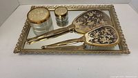 Vintage mirror tray 15x10 inches with ornate gold-toned frame, paired with matching hand mirror and hairbrush with floral designs, plus two glass lidded bowls with fabric and gold rim lids.