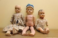 Three antique dolls posed sitting against a wall showing overall condition and clothing.