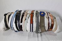 Photo showing 15 belts arranged horizontally on a white pillow, featuring various materials and buckle styles.