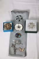 Overview of all eight brooches arranged on a grey cloth, showing variety of styles and materials including Indian silver, mother of pearl, stones and crystals.