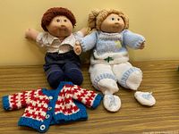 Both Cabbage Patch Kids dolls, Nathanial and Marcella, sitting side by side with their outfits visible on a wooden surface against a yellow wall. Extra hand-knit sweater laid in front.