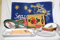 View of the entire lot showing blue Christmas doormat reading 'Season', red and white Christmas plates, packaged tablecloth, cake plate with server, woven basket, trays and bowls.