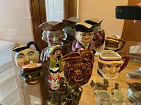 Overview of 8 ceramic character jugs with faces displayed on glass shelf