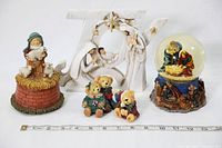 Photo showing all items including carved wooden JOY nativity, snow globe nativity, music box with child and lambs, and three teddy bear figurines, ruler for scale.