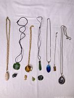 Image showing 7 necklaces with various colored stone pendants and 5 loose small pendants arranged on white fabric.
