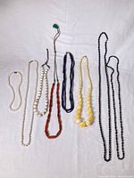 Eight vintage bead necklaces displayed on white cloth background showing various bead styles and colors.