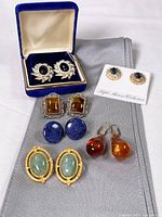 Photo showing 6 pairs of pierced earrings on a grey cloth: black stone with rhinestones in blue velvet box, gold-tone teal stone rhinestone pair on card, square amber stones, large blue oval stones, green oval stones in gold-tone setting, amber teardrop earrings