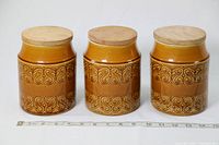 Set of three brown glazed ceramic canisters with embossed decorative bands and wooden lids, lined up side by side on a white background with ruler for scale