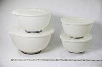 Four white plastic mixing bowls with their matching lids arranged side by side by size on a white background, with a ruler for scale.