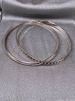 Two thin silver bracelets, one plain and one decorated, on a silver-gray fabric background.