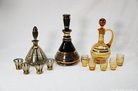 Full set of three vintage MCM style glass decanters and matching glasses in amber and black with gold accents arranged in one photo.