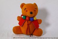 Front view of the vintage orange plush bear wearing a colorful beaded necklace and red felt vest, sitting on a white background with measuring tape visible.