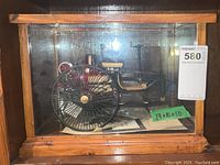 Overall shot of model inside wood and glass display case, showing large rear spoked wheel and documents below.