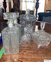 Group of five crystal items on wooden table including two decanters, candy dish, bell, and a large covered jar.