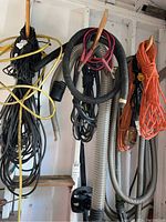 Photo showing various extension cords, corrugated hoses, black hoses, jumper cables, and orange netted rope hanging on a wall.