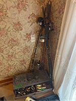 Fishing rods and reels standing upright beside a floral wallpapered wall near a window. Metal tackle box with open lids showing fishing lures inside placed on the floor.