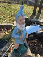 Concrete garden gnome with blue hat and tunic, white beard, standing on green base. Paint shows wear and chipping.