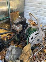 Gas powered garden tiller next to house with garden hose and hose caddy, showing overall condition and outdoor setting.