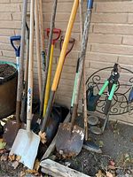 Various long-handled garden tools including shovels, spades, snow shovel, and rakes leaning against a brick wall with visible dirt and signs of use.