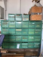 Full front view of green metal cabinet with yellow and white handwritten labels indicating hardware contents in each of 24 drawers, plus 3 extra metal drawers and wood boxes on top.