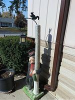 Outdoor concrete statue of a man wearing a hat and scarf, holding a tall lamppost painted cream with a hanging black lantern and a squirrel decoration on top.