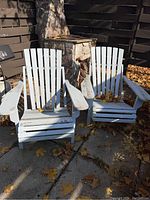 Pair of white painted wooden Muskoka chairs placed outdoors on a concrete surface surrounded by fallen leaves, showing wear and paint chipping.