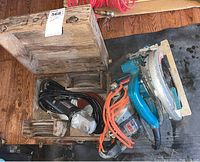 Photo showing Makita magnesium circular saw with attached power cord, Craftsman angle grinder and Skil Xtra tool in a wooden storage box on floor.