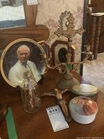 Photo of the lot showing a crucifix candle holder, glass bottle with miniature Calvary scene, vanity boxes, and papal plates arranged on a wooden surface.