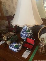 Photo of blue and white porcelain table lamp with floral patterns, white fabric lampshade, and nearby decorative items including carved elephant figures, a small blue and white vanity box, and a small red enameled heart-shaped box open inside a red box.