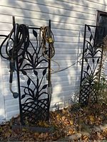 Four black metal garden trellises leaning against a white exterior wall, featuring decorative leaf design and some garden tools hanging on them.