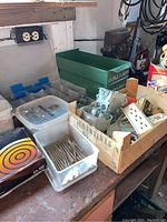 Overview of workbench surface showing hardware bins, wooden crate with electrical parts, plastic containers with screws and nails, metal tin with target design