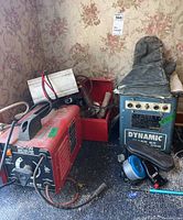 AC-100 Arc Welder next to Dynamic Mark 88 Welder, toolbox, gloves, and accessories on floor with floral wallpaper background