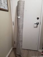Rolled large light gray rug standing upright near a white door, showing tag with size information.