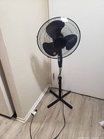 Full view of the electric pedestal fan showing the three black blades, circular metal grille, adjustable height stand with cross base, and power cord plugged into a wall outlet.