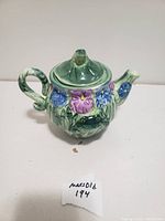 Front view of glass frog teapot showing frog detail and floral decoration on body with attached handle and spout and closed lid.