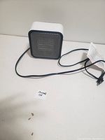Small cube-shaped electric heater with white body and black front grille, power cord and attached paper tag, photographed on a flat surface.