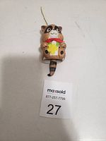 Front view of the raccoon hanging bell ornament showing facial details, flower, and hanging string