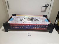 Top angled view of the air hockey table showing NHL rink surface, various NHL team logos around the sides, included accessories in plastic bag on top
