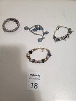 Four charm bracelets displayed on a white surface: two silver-toned, one with blue stones, one gold-toned with black and gold beads.