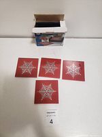 Four square red coasters with a white snowflake design displayed in front of the open box.
