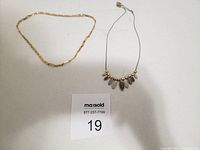 Two custom necklaces displayed on a white background. One gold-tone chain with a Greek key pattern and one silver-tone wire chain with multiple oval pendants.