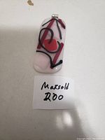 Photo shows a rectangular fused glass pendant with a white background and abstract red and black lines and shapes. The pendant is photographed on a white surface next to a small paper note with 'Maxsold 200' written on it.