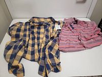 Two men's medium size long-sleeve shirts laid flat. Left shirt is yellow and navy blue plaid button-up. Right shirt is pink, orange, and navy striped polo.