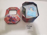 Photo of Pokemon cards inside a red Garchomp metal storage tin, cards stacked inside the open tin next to its lid.
